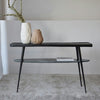 La Consola Lena Metal Negro Mármol Verde es una mesa consola minimalista negra con cuatro patas delgadas, que cuenta con un estante superior adornado con jarrones y un estante inferior que sostiene un libro. Su diseño elegante complementa la decoración contemporánea sobre un fondo claro, realzando la elegancia del espacio.