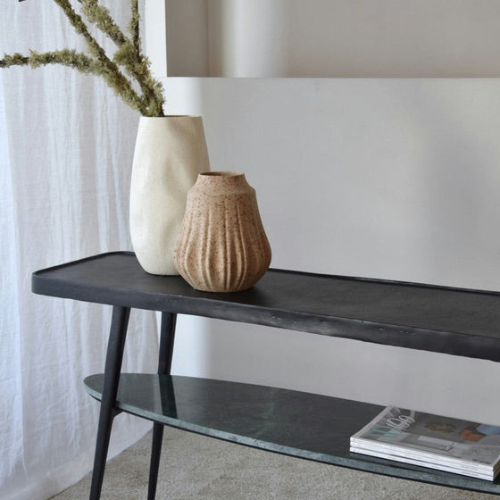 Dos jarrones de cerámica, uno blanco y otro marrón, adornan la elegante Consola Lena Metal Negro Mármol Verde junto a una cortina blanca. Las revistas descansan en el estante de vidrio inferior, lo que ofrece un toque de decoración contemporáneo. Las ramas secas llenan elegantemente el jarrón blanco.