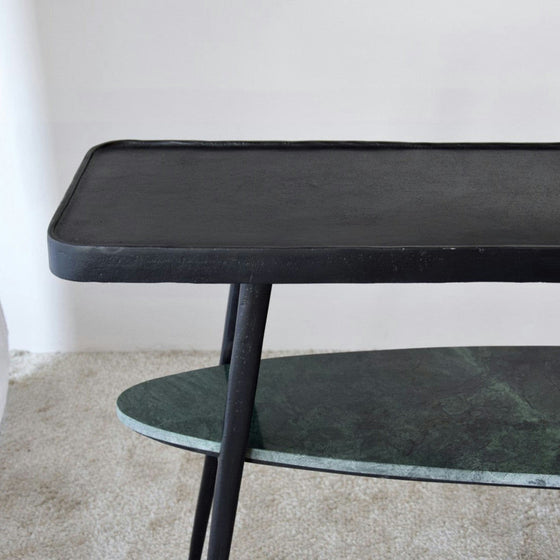 Un primer plano de la Consola Lena Metal Negro Mármol Verde, una mesa auxiliar de dos niveles de metal negro con líneas elegantes y una parte superior rectangular y un estante inferior ovalado, colocada sobre una alfombra clara, realza cualquier espacio con decoración contemporánea.