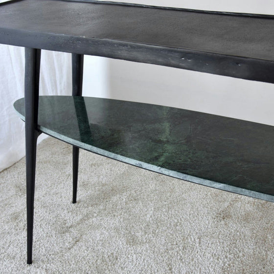 La Consola Lena Metal Negro Mármol Verde es una mesa negra de dos niveles que presenta un tablero rectangular delgado y un estante inferior de mármol verde, que se coloca elegantemente sobre un piso alfombrado y encarna una decoración contemporánea.