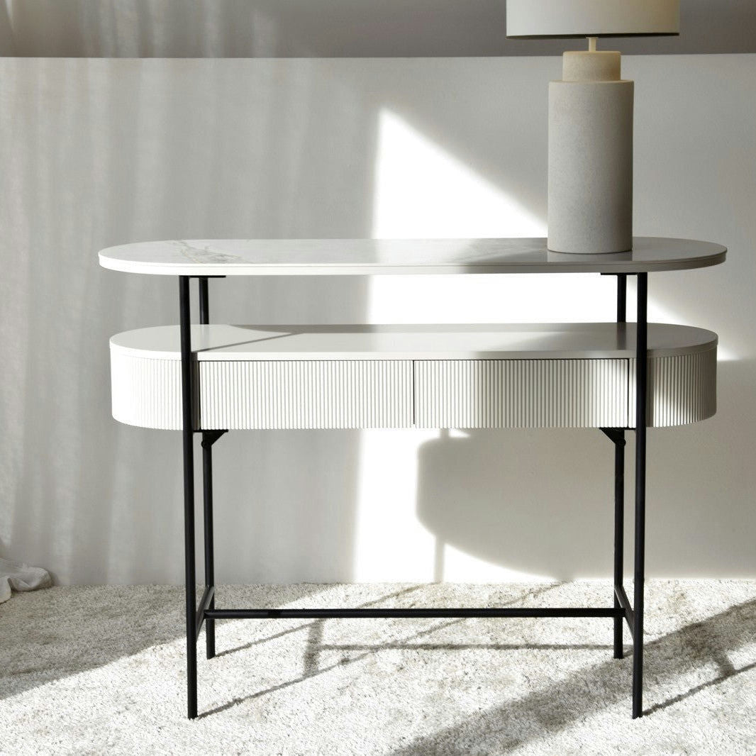La mesa Consola Oval N.24, en Gris Arena con tablero de cerámica blanca y gris, se yergue elegante sobre una alfombra clara. Sostiene una lámpara cilíndrica con pantalla blanca, mientras la luz del sol crea sombras artísticas en la pared, realzando la decoración y el interiorismo.