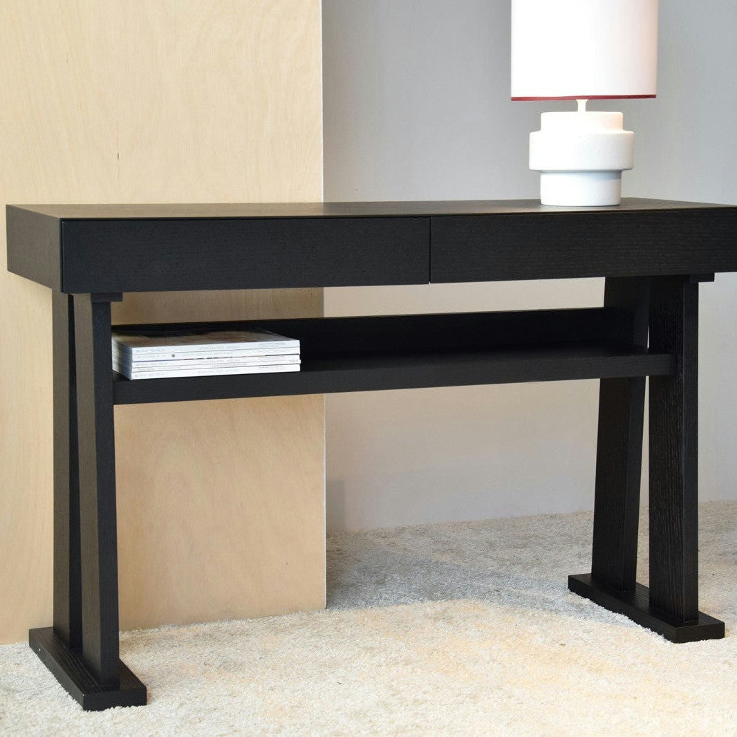 Una elegante Consola Roble Negro Mate - Estilo Loft con dos cajones y un estante inferior de libros, rematado por una lámpara blanca. Su diseño minimalista complementa a la perfección la alfombra de color claro.