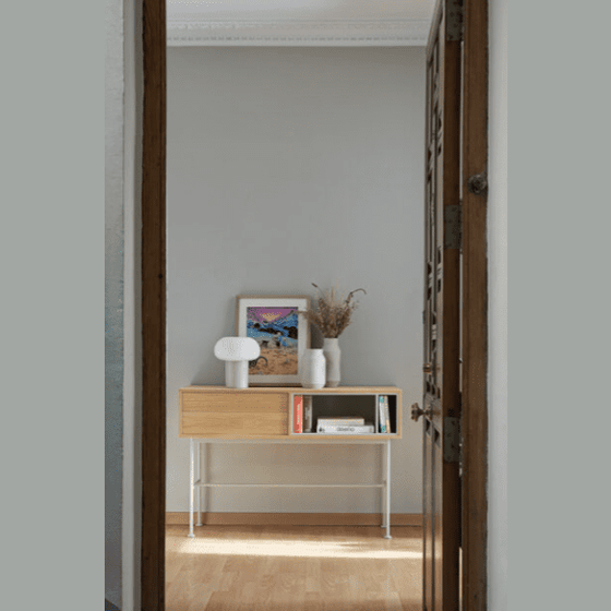 Una pequeña Consola Yoko 2C de madera en un rincón, adornada con una lámpara, un cuadro enmarcado, libros y un jarrón con flores secas, vista desde una puerta.