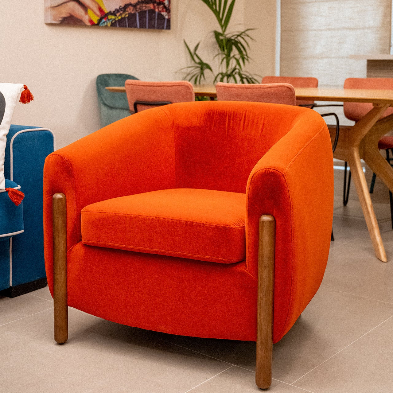 El Sillón Tapizado en Rojo Nest, un sillón naranja vibrante con patas de madera elaboradas con madera de caucho duradera, se encuentra en un interior moderno junto a una mesa de comedor y sillas coloridas.
