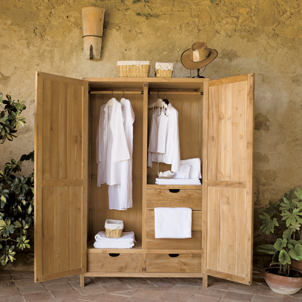 El Armario en Madera Teca Rústico Marco, que exhibe camisas blancas, toallas dobladas y canastas de mimbre en la parte superior para brindar diseño y practicidad. Un sombrero reposa en el borde de esta elegante pieza elaborada con madera de teca duradera.