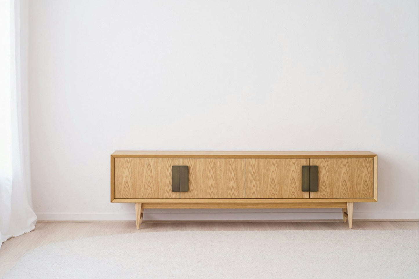 El Mueble TV en DM y roble Oia es un aparador minimalista con cuatro puertas con tiradores negros y un diseño elegante. Fabricado en roble, se destaca elegantemente contra una pared blanca sobre un piso de madera clara.