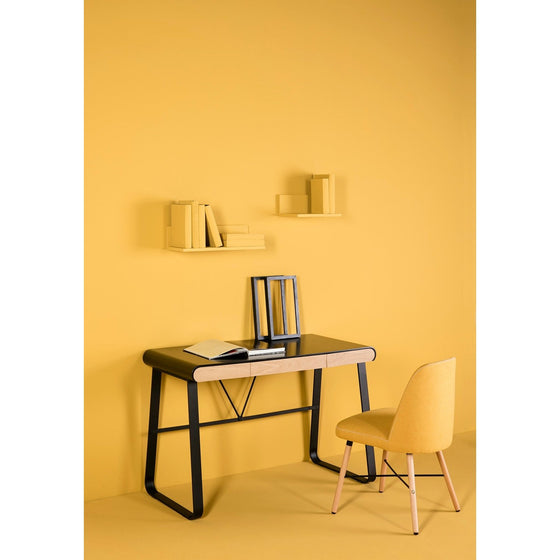 Una habitación de paredes amarillas con un Escritorio de Metal con cajones Astrid negro, un libro y un objeto enmarcado. En la pared hay dos pequeños estantes con algunos libros. Una silla amarilla se coloca frente al escritorio, creando una oficina moderna con sutiles toques de estilo industrial.