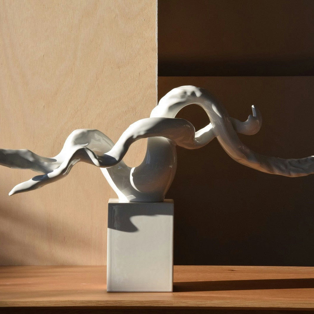 La Escultura Abstracta Rama Cerámica Blanco - Elegancia Zen, con sus formas retorcidas sobre una elegante base cuadrada, encarna el arte contemporáneo al proyectar una sombra sobre una superficie de madera.