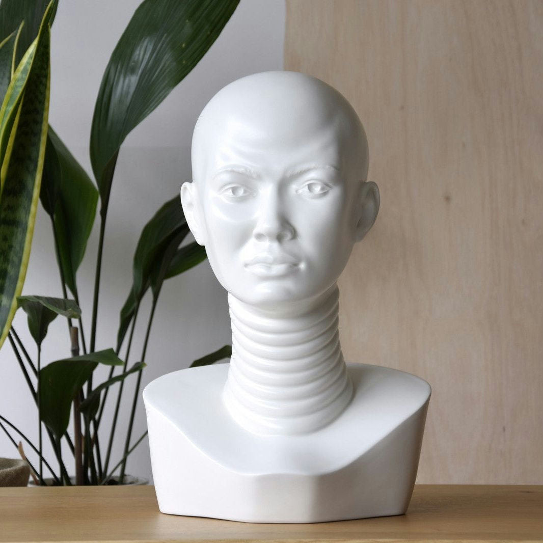 La Escultura África en cerámica Biscuit Matte, que representa una cabeza humana calva con un cuello alargado y segmentado, reposa sobre una superficie de madera rodeada de frondosas plantas. Es la pieza perfecta para añadir un toque artístico a la decoración de interiores.