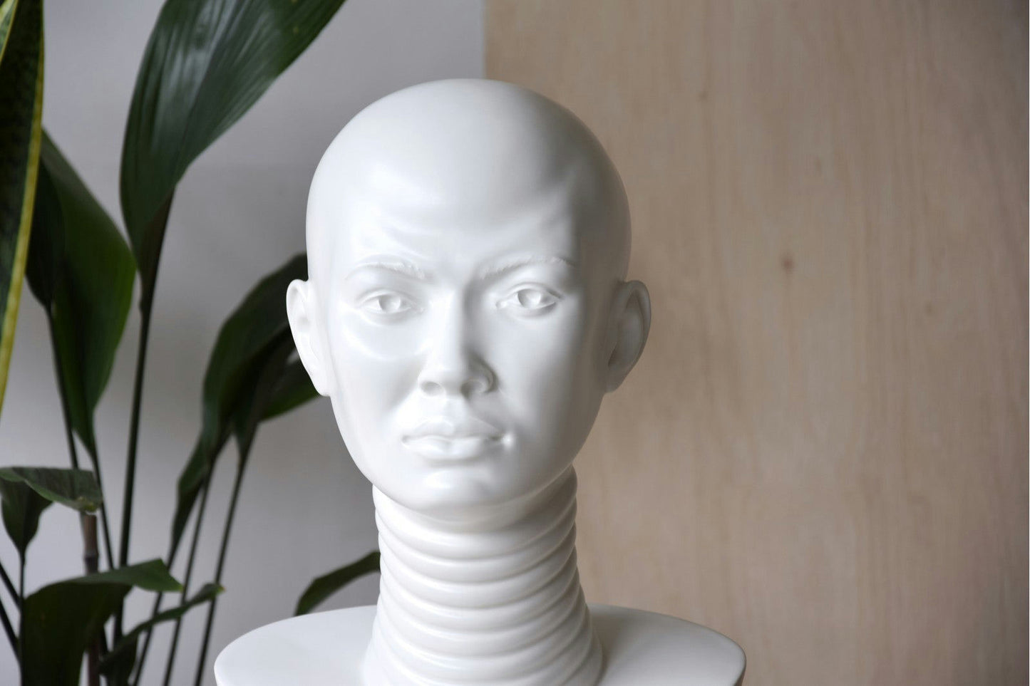 La Escultura África en cerámica blanca Biscuit Mate, con un cuello alargado, se destaca sobre un fondo de madera con hojas verdes a la izquierda. Muestra un diseño artístico africano único, que agrega un toque exótico a la decoración de interiores.