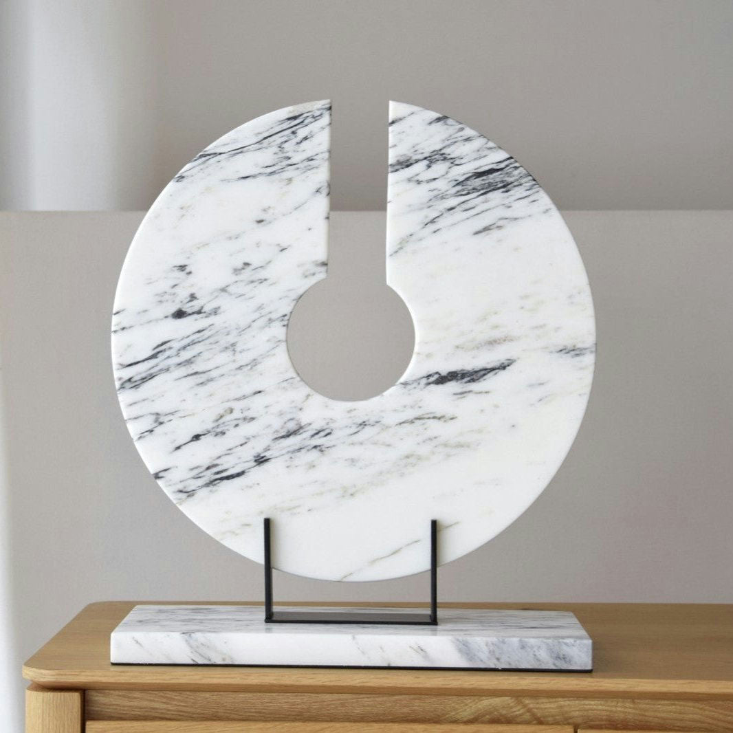 La Escultura Arnela N2, realizada en mármol y metal, presenta un diseño circular con un hueco vertical y reposa sobre una base rectangular. Su diseño sofisticado la hace ideal para la decoración de interiores, realzando bellamente cualquier superficie de madera.