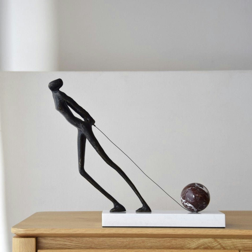 La Escultura Arrastre presenta una esbelta figura humana tirando de una piedra redonda y oscura sobre una superficie de madera, realzando la decoración de interiores con su elegante diseño de metal y mármol.