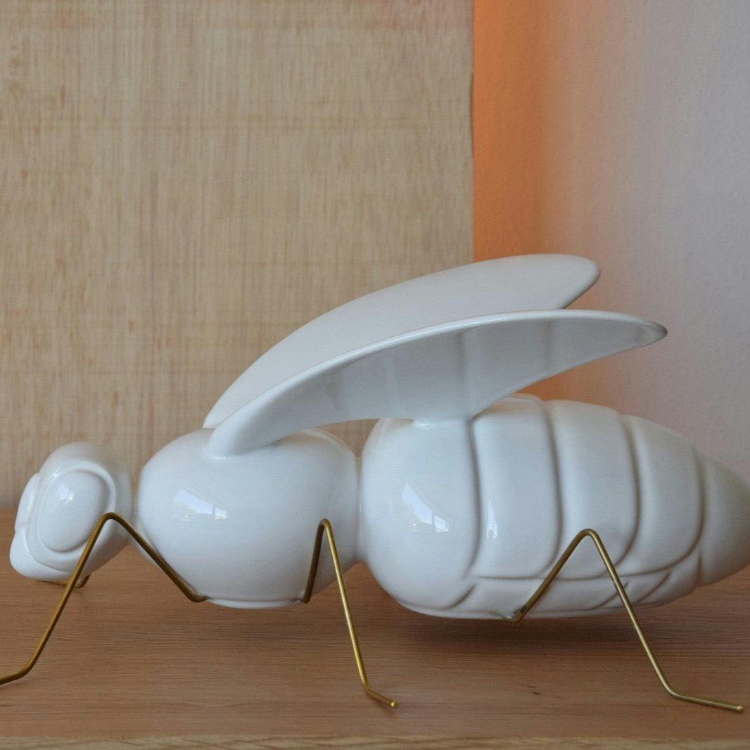 La Escultura Cerámica Abella Blanco Brillo muestra una abeja de cerámica blanca con grandes alas y patas doradas, elegantemente colocada sobre madera. Esta pieza encarna la decoración interior elegante y agrega sofisticación a cualquier espacio.