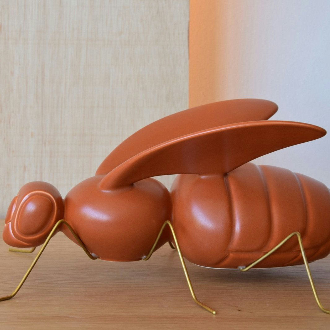 La Escultura Cerámica Abella Teja Mate es una gran escultura de hormiga abstracta de color marrón con rasgos exagerados y patas de metal sobre una superficie de madera, que complementa perfectamente cualquier decoración de interiores.