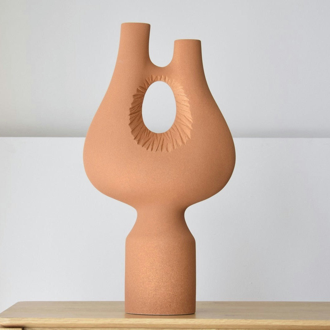 Un jarrón de cerámica beige de la Escultura Cerámica Artesanal Colección Vacío, Pieza 1, Textura Teja, con dos caños y una abertura circular texturizada en el centro, se encuentra sobre una superficie de madera sobre un fondo liso.