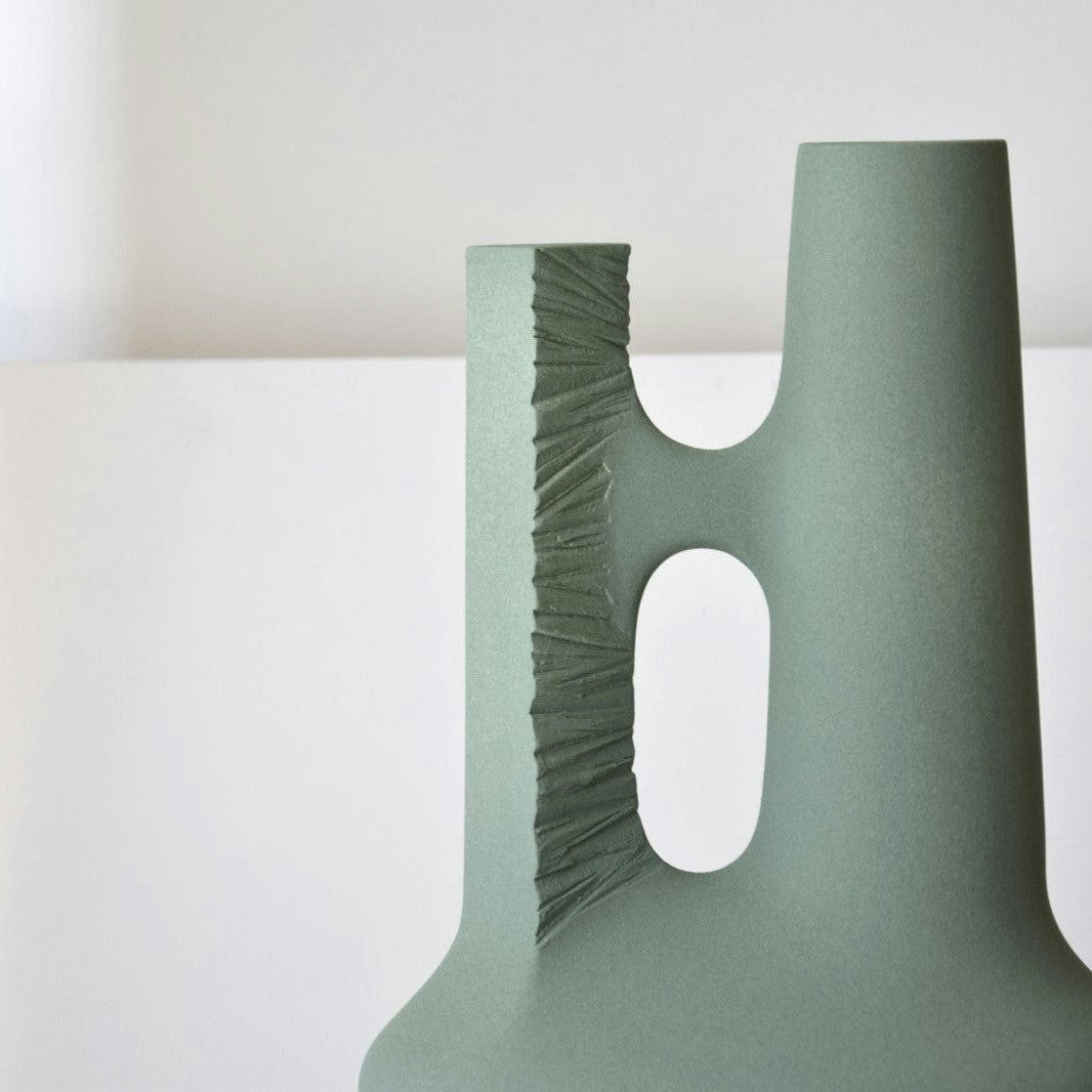 La Escultura Cerámica Artesanal Colección Vacío: Pieza 2 presenta un jarrón de cerámica verde con una ranura vertical texturizada de inspiración natural de la erosión y dos aberturas de diferentes tamaños, todas expuestas sobre un fondo blanco liso.