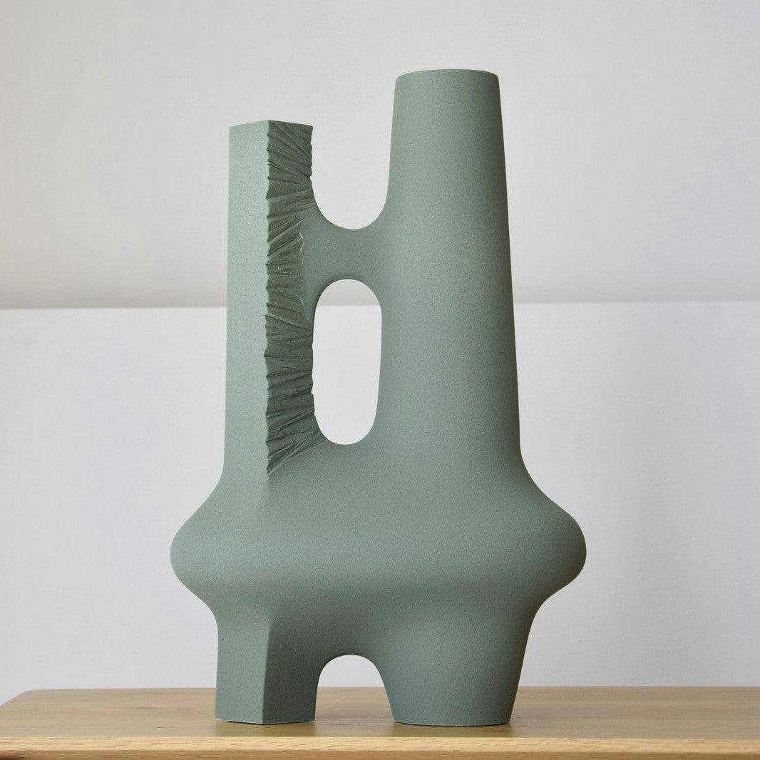 La "Escultura Cerámica Artesanal Colección Vacío. Pieza 2. Textura Verde" es una moderna escultura de cerámica verde hecha a mano con superficies lisas y texturizadas, que presenta formas geométricas abstractas sobre una mesa de madera sobre un fondo gris claro.