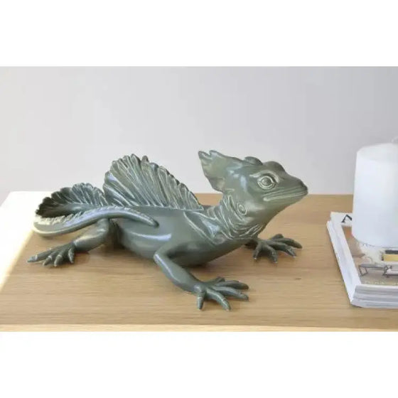 Una Escultura Cerámica Basilisco Verde de la Colección Artesanía Natural se encuentra sobre una mesa junto a una vela, agregando un toque de artesanía natural a cualquier hogar.