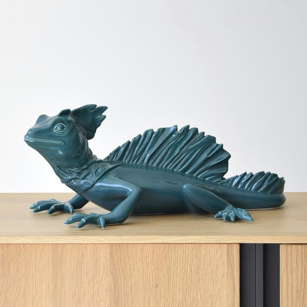 La Escultura Cerámica Basilisco Verde Brillante presenta un lagarto verde azulado con un lomo puntiagudo y detalles intrincados, exhibido sobre una superficie de madera sobre un fondo simple.