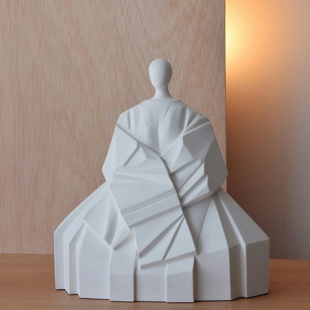 Una escultura geométrica blanca llamada "Escultura Cerámica Japonesa Manga Extendida Blanco Mate" representa una figura sentada con ropa envuelta sobre un fondo de madera clara. Rezuma decoración zen, realzada por una suave iluminación cálida desde la derecha.