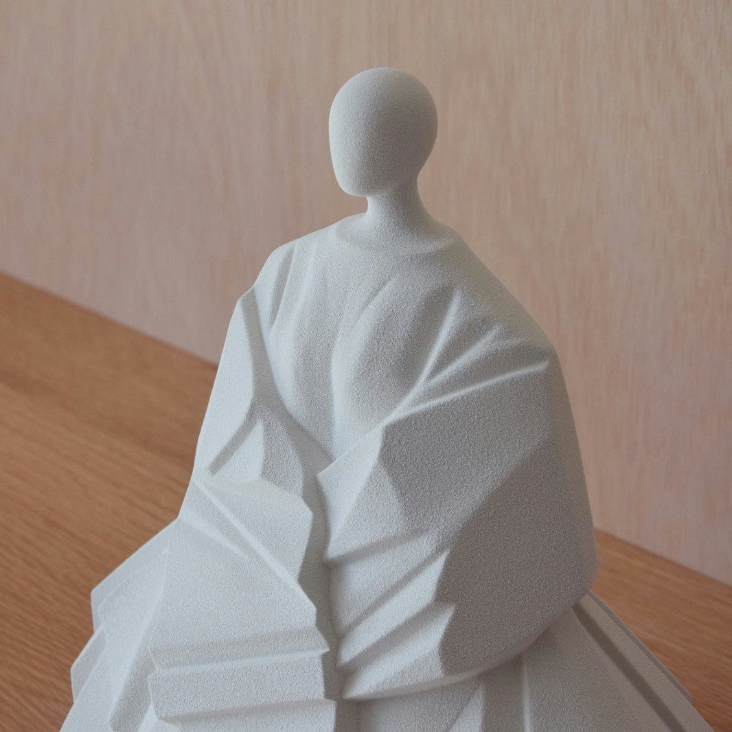 La "Escultura Cerámica Japonesa Manga Extendida Blanco Mate" es una escultura abstracta de una figura sin rostro con patrones geométricos, que se asemeja a una tela drapeada sobre una superficie de madera. Rezuma la calma y sencillez de Decoración Zen, como una delicada figura de cerámica japonesa.