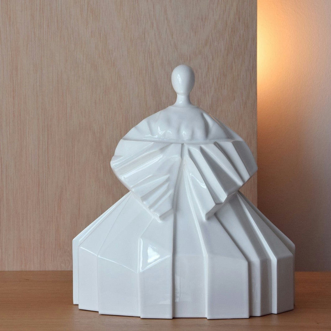 La Escultura Cerámica Japonesa Manga Recogida Blanco Brillo exhibe un diseño minimalista con vestido geométrico. Está sobre una superficie de madera, con fondo de madera y una iluminación suave que acentúa su serenidad Zen.