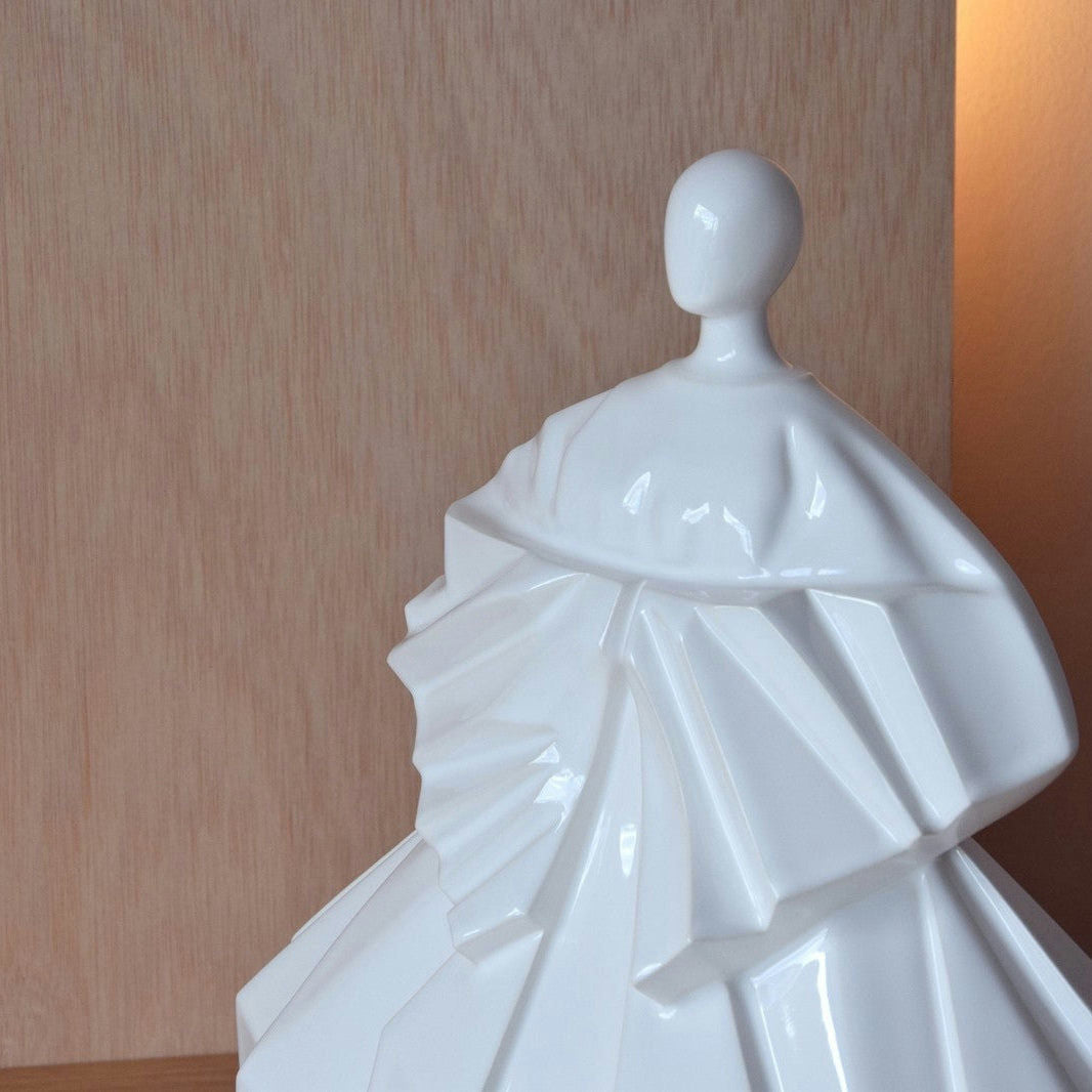 La Escultura Cerámica Japonesa Manga Recogida Blanco Brillo presenta una figura sin rostro con una prenda fluida y con volantes con simplicidad zen. Elegantemente colocado sobre un fondo de madera, proporciona una estética serena para la decoración de interiores.
