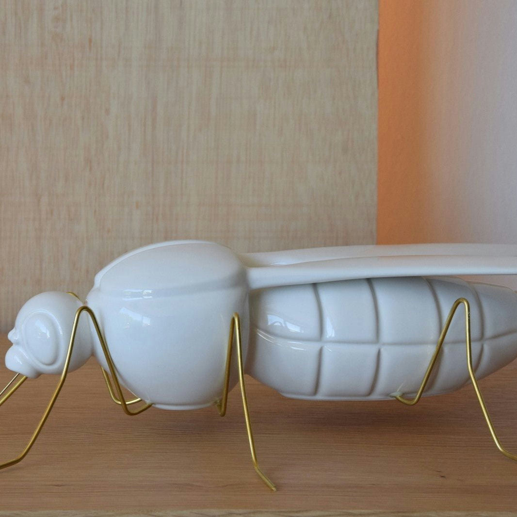 La Escultura Cerámica Mosquito Blanco Brillo, con patas de alambre dorado, irradia elegancia dorada sobre una superficie de madera contra una pared de color claro.