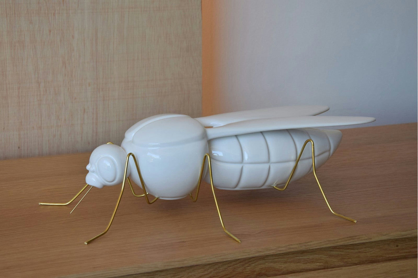 La Escultura Cerámica Mosquito Blanco Brillo presenta un cuerpo blanco brillante y delgadas patas y antenas de color dorado metálico, emanando un aura de elegancia dorada mientras descansa elegantemente sobre una superficie de madera clara.