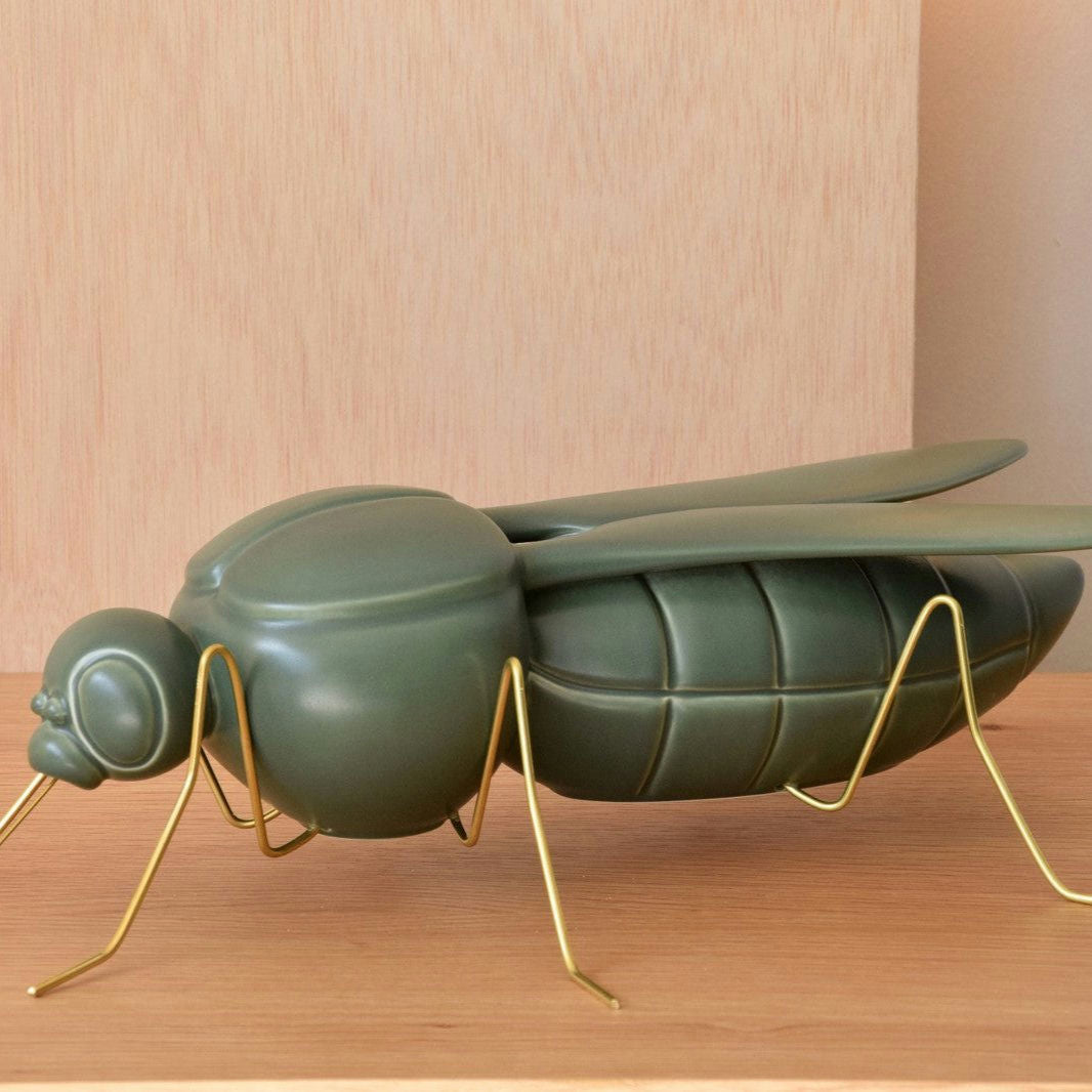 La Escultura Cerámica Mosquito Verde Militar Mate, un gran modelo de mosca de cerámica verde con patas de alambre dorado, adorna la superficie de madera. Su encanto único y su acabado verde militar mate realzan la decoración y cautivan a los admiradores del arte cerámico.