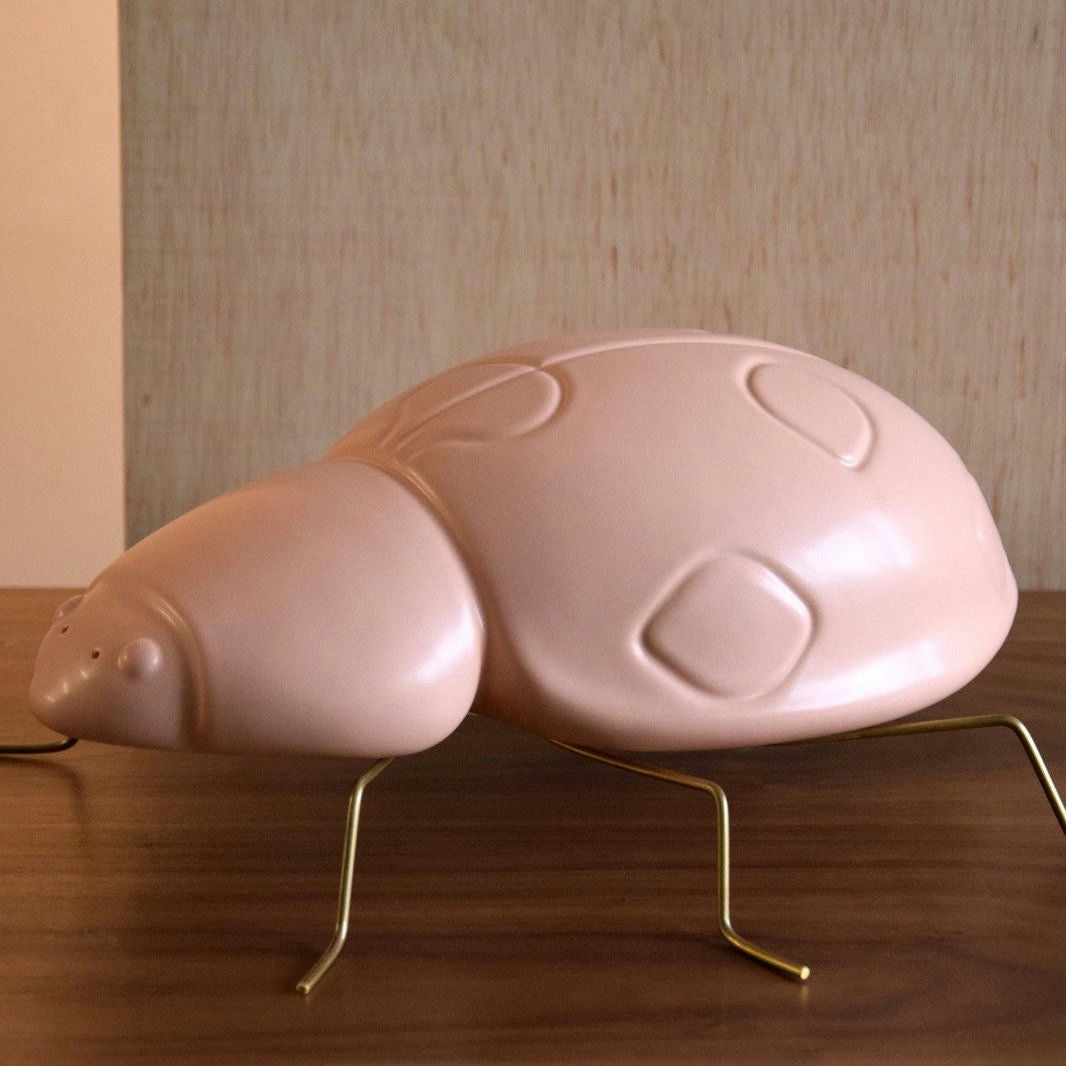 La Escultura Cerámica Xoanina Rosa Mate, una gran escultura rosa lisa con patas de alambre dorado, se muestra sobre una superficie de madera sobre un fondo neutro, perfecto para la decoración de interiores.