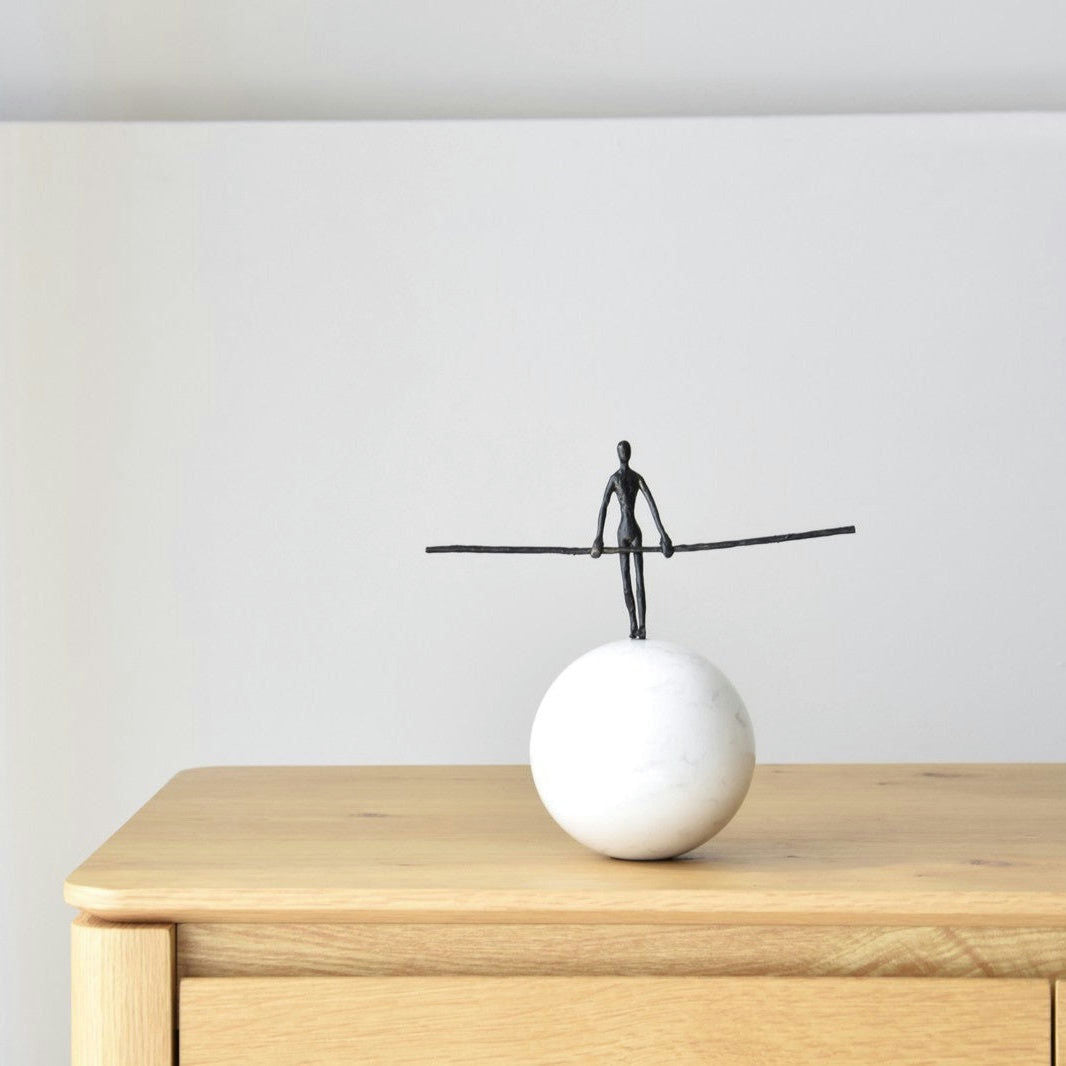 La Escultura Funambulista N1: Metal y Mármol es una pequeña escultura de metal de una figura en equilibrio sobre una base esférica blanca con un poste extendido, ideal para decoración de interiores y elegantemente colocada sobre una superficie de madera.