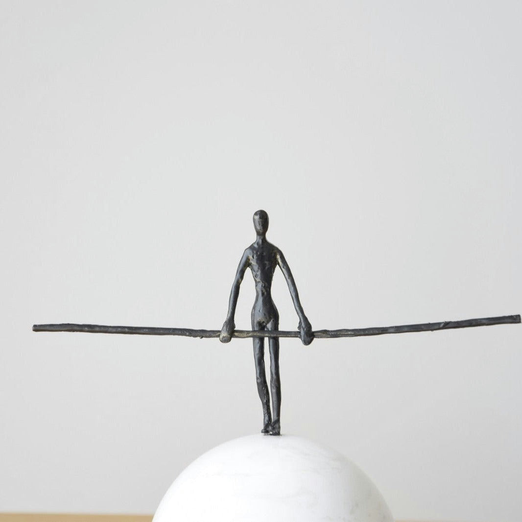 La Escultura Funambulista N1: Metal y Mármol es una pequeña escultura de metal de una figura que se balancea elegantemente sobre un alambre delgado encima de una base esférica blanca y lisa, perfecta para agregar un toque elegante a cualquier decoración interior.