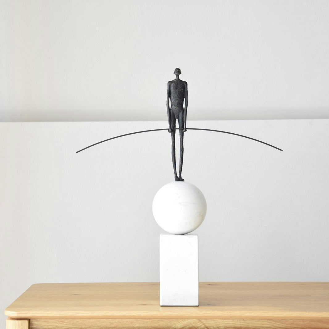 La Escultura Funambulista N2: Metal y Mármol presenta un diseño único y atemporal que presenta una pequeña figura en delicado equilibrio sobre una esfera blanca en un bloque rectangular, sosteniendo con gracia un poste largo y delgado.