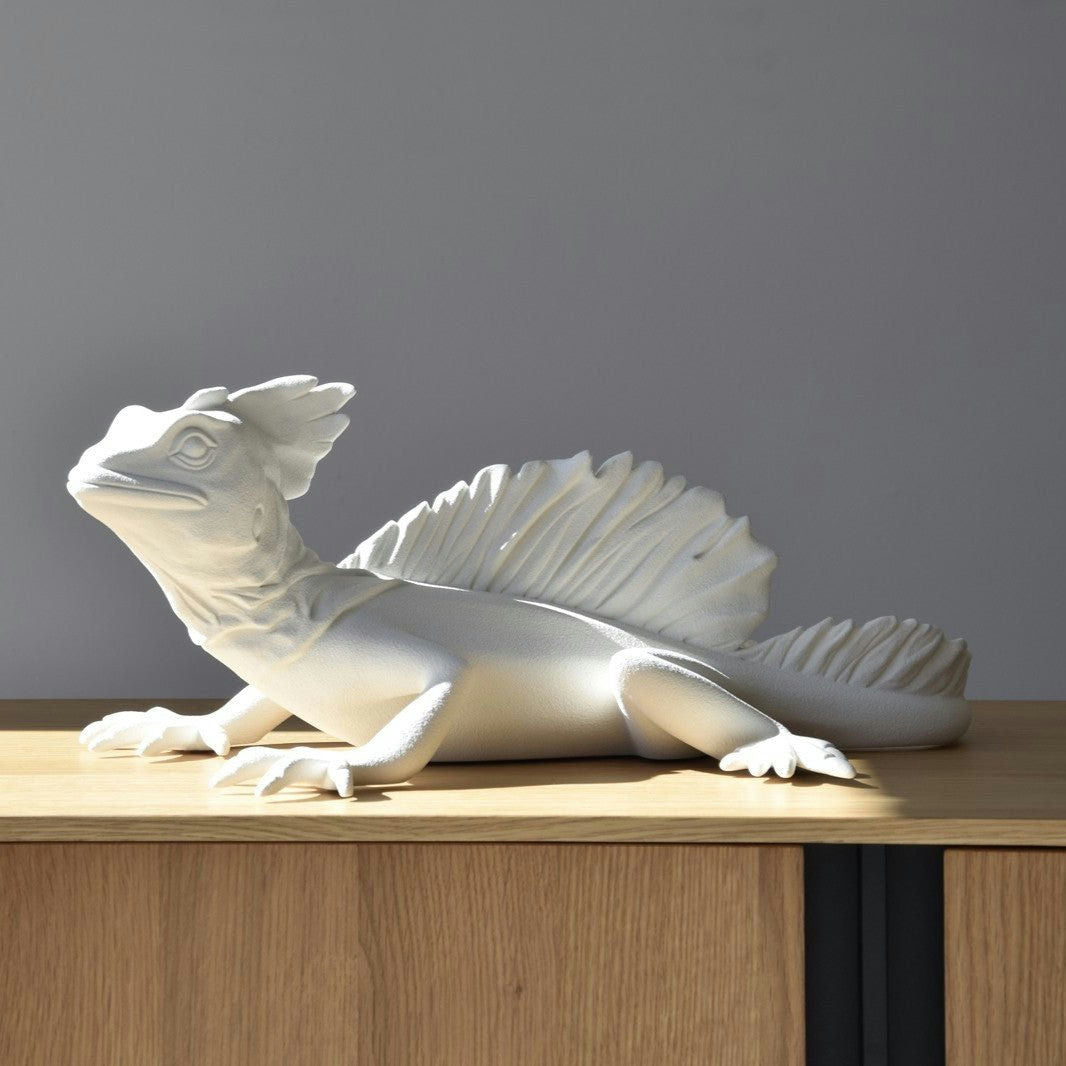 La Escultura de Cerámica Basilisco Blanco Mate es una escultura de cerámica blanca de un lagarto con escamas y aletas texturizadas, que se exhibe sobre una superficie de madera contra un fondo gris. Forma parte de la Colección Arte Artesanal, que destaca la artesanía y los detalles intrincados.