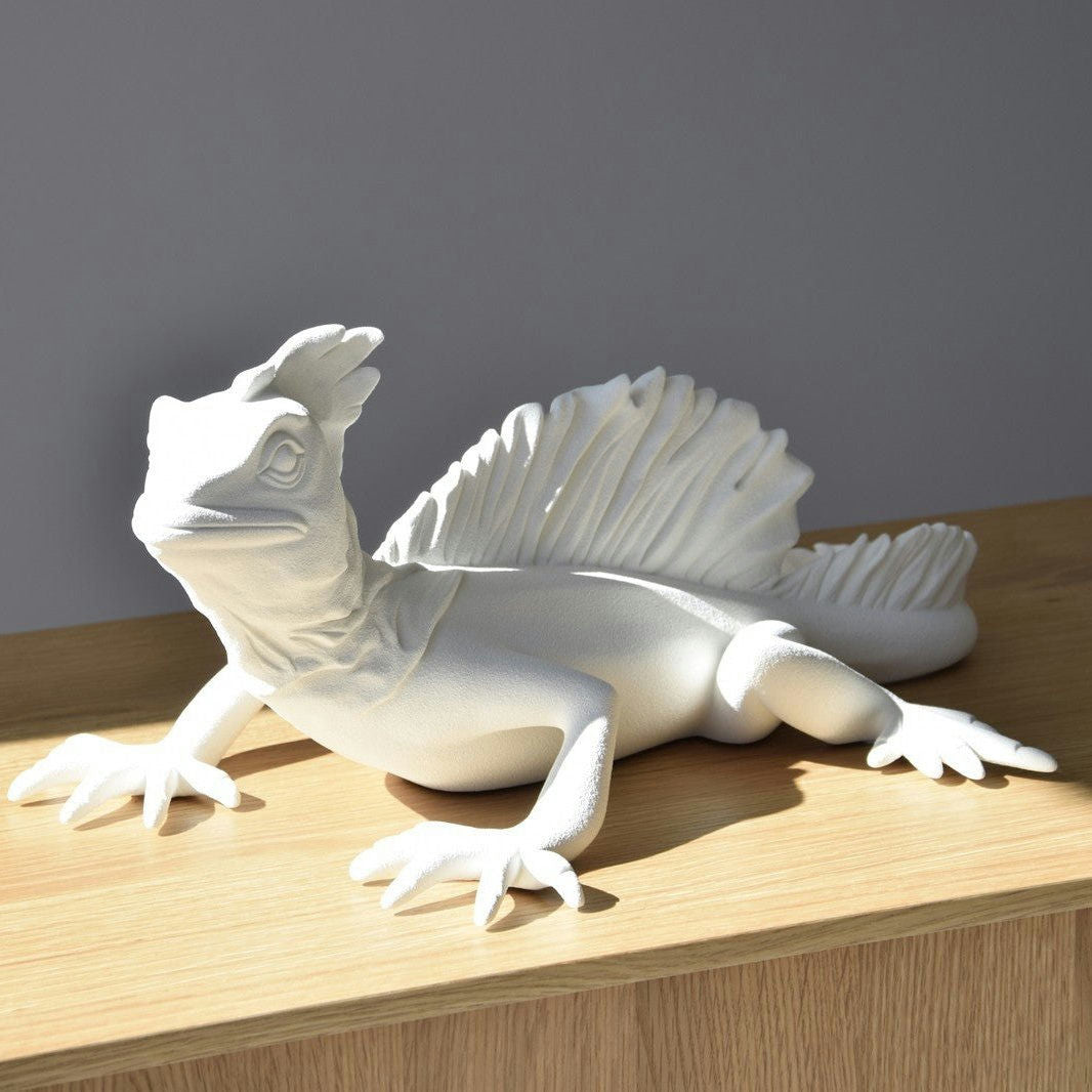 Una detallada Escultura de Cerámica Basilisco Blanco Mate con un respaldo con volantes se encuentra sobre una superficie de madera, iluminada por la luz del sol contra un fondo gris; parte de la exquisita Colección Arte Artesanal.