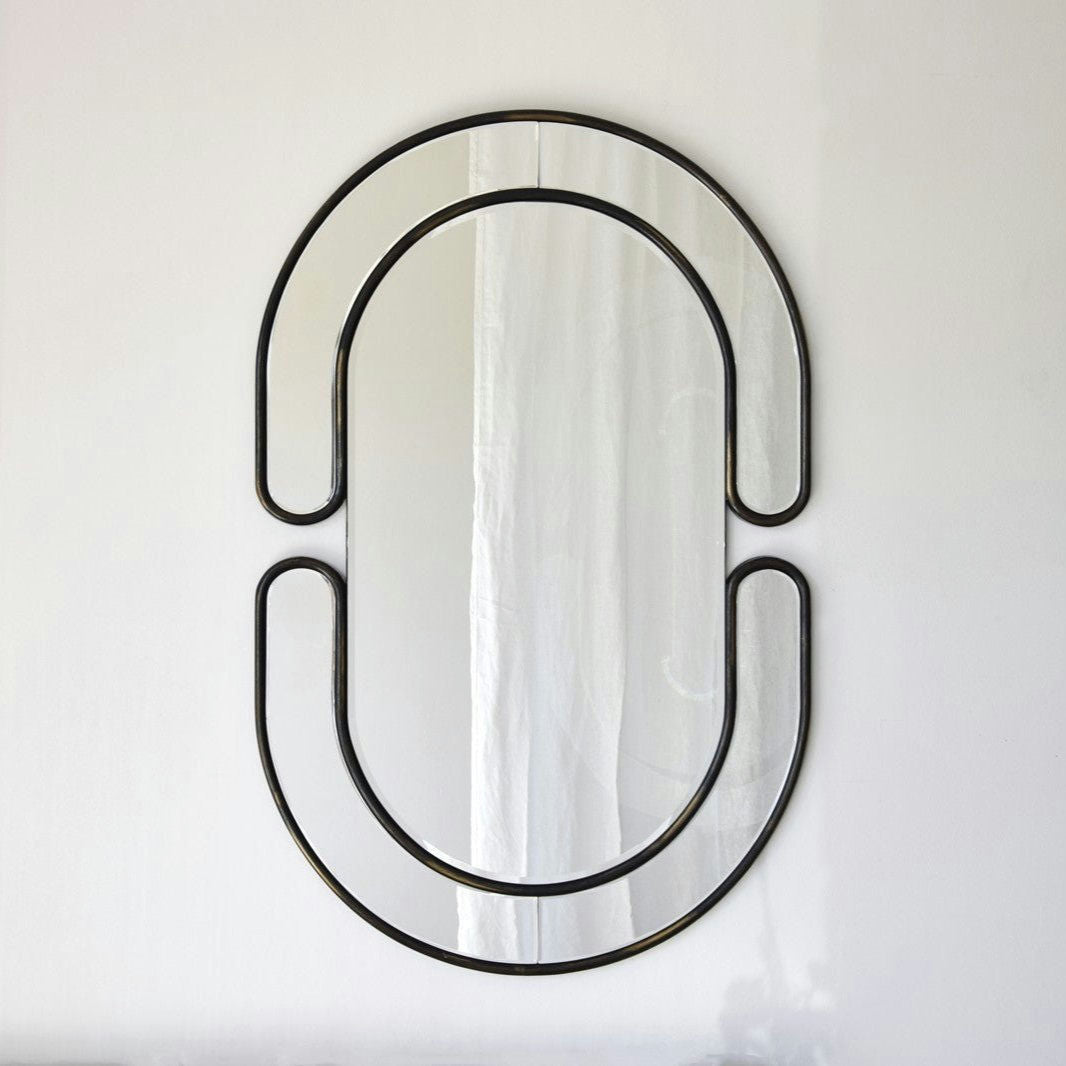 El Espejo Decorativo Oval N.1, con marco negro y líneas curvas entrelazadas simétricas, cuelga elegantemente en una pared blanca, realzando cualquier diseño interior.
