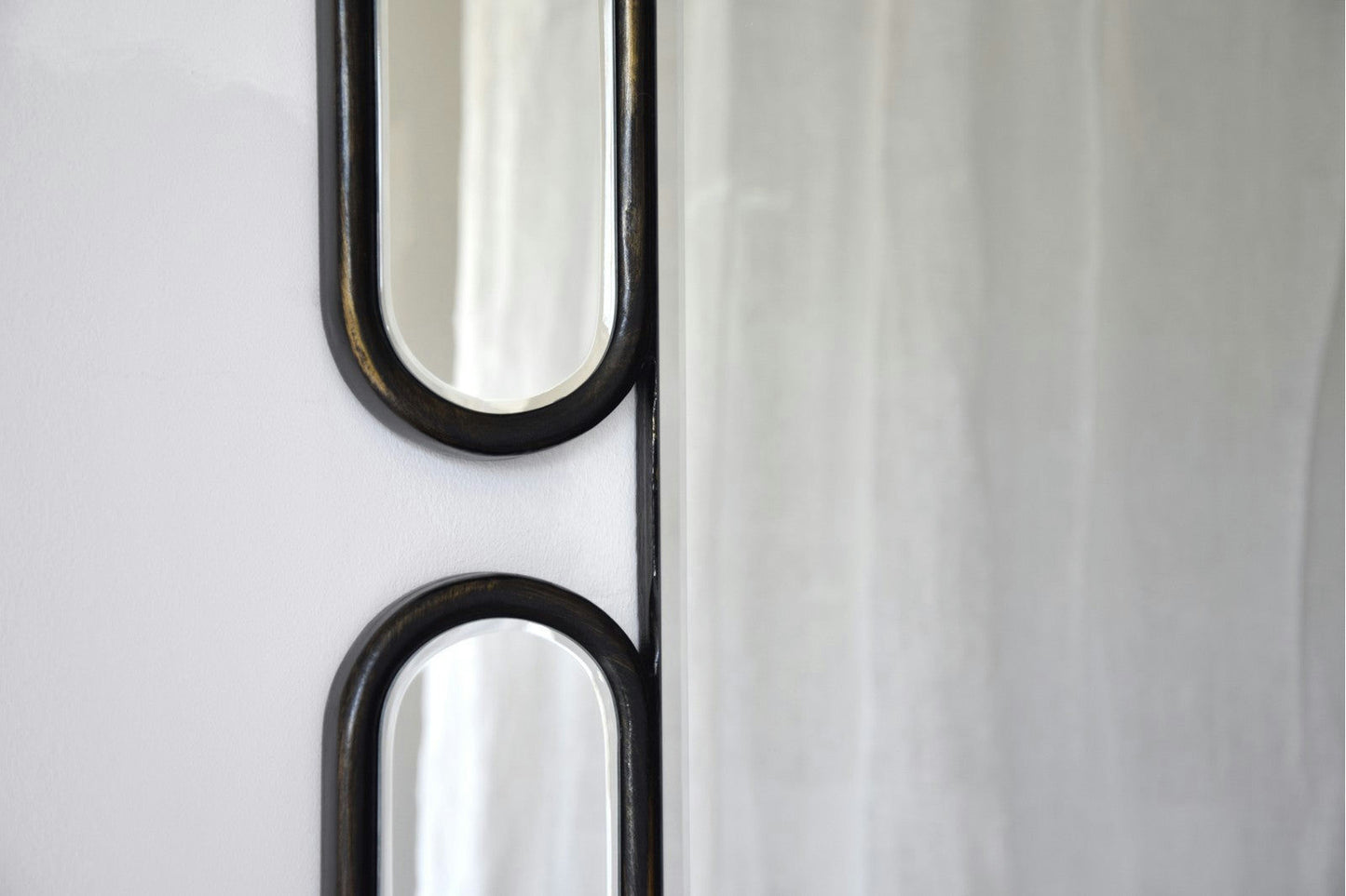 Primer plano del Espejo Decorativo Oval N.1, con un elegante marco de MDF negro, montado con elegancia sobre una pared blanca. El espejo refleja una cortina blanca suavemente difuminada, lo que le aporta un toque elegante al diseño interior.