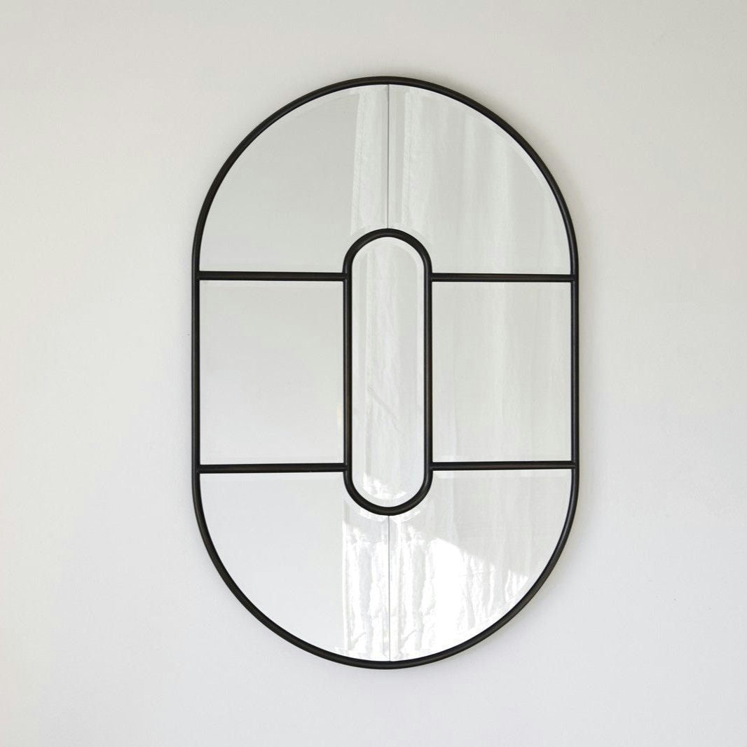 El Espejo Decorativo Oval N.2, dividido por un fino marco negro, refleja una pared blanca lisa y encarna el diseño contemporáneo.