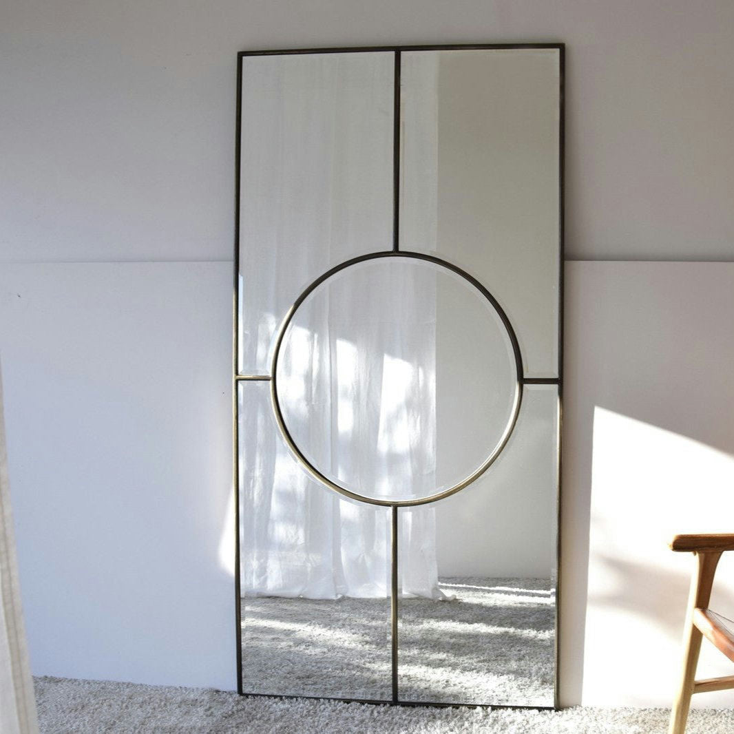 El Espejo Decorativo de Suelo - Circulo, con un diseño geométrico, se apoya contra una pared blanca sobre un suelo alfombrado. Una silla de madera añade un toque de interiorismo moderno al espacio.