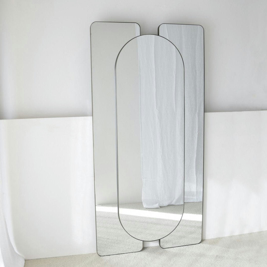 El Espejo Decorativo de Suelo Metal Oscuro, un diseño contemporáneo independiente de forma ovalada y rectangular, se apoya con estilo contra una pared blanca y un piso alfombrado.