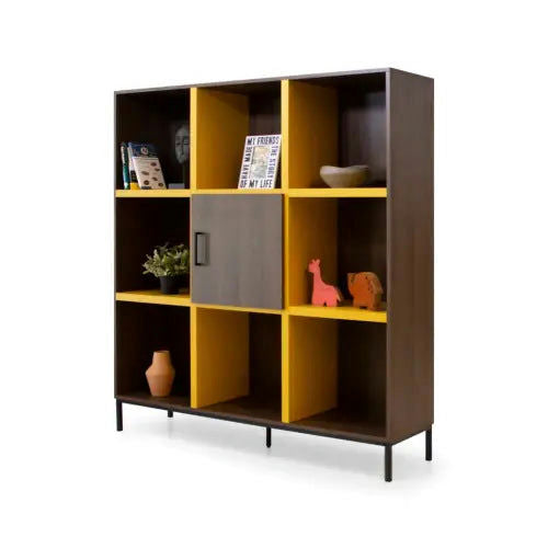 La Estantería Dinka 02 en Amarillo es una estantería de madera con compartimentos abiertos y cerrados, adornada con libros, jarrones y figuras. Cuenta con un marco marrón oscuro y un respaldo amarillo, elaborado a partir de elegante madera de nogal para un diseño contemporáneo.