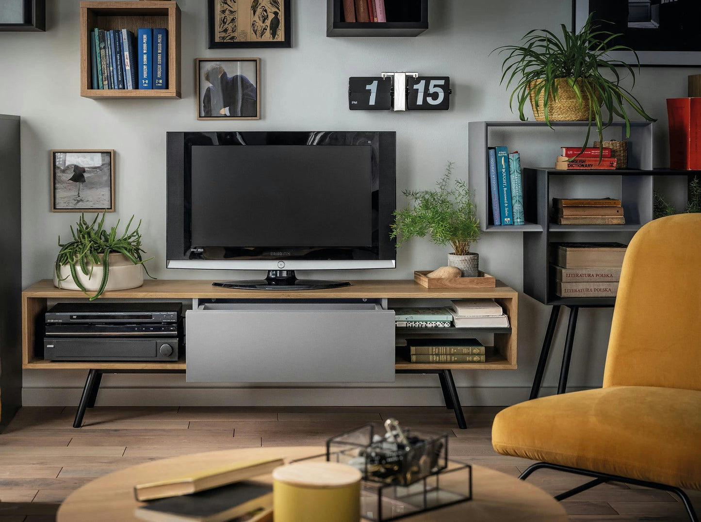 Una sala de estar moderna muestra un televisor sobre una consola de madera, rodeado de plantas, libros y una estantería geométrica Kuadrat en Roble, Grafito y Gris. Una silla de color amarillo mostaza encarna el diseño contemporáneo en el frente, mientras que un reloj de pared marca tranquilamente la 1:15.