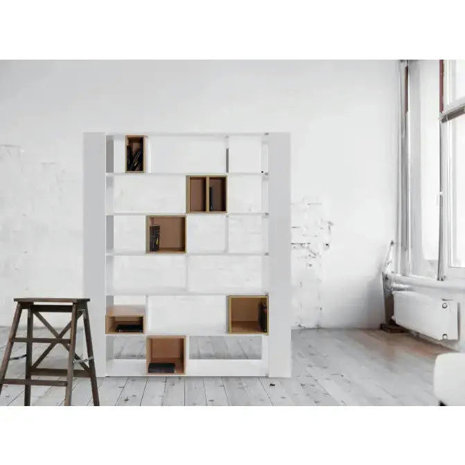 La Estantería Vooxy Libreria 01, una moderna estantería blanca de ecomadera con múltiples compartimentos, algunos llenos de libros, se encuentra en una habitación minimalista con paredes blancas y piso de madera. Un taburete de madera cercano realza el diseño de interiores.