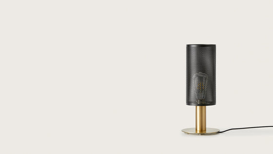 Una moderna lámpara de mesa Fito cilíndrica de malla negra con base dorada, situada a la derecha contra un fondo claro y sencillo.