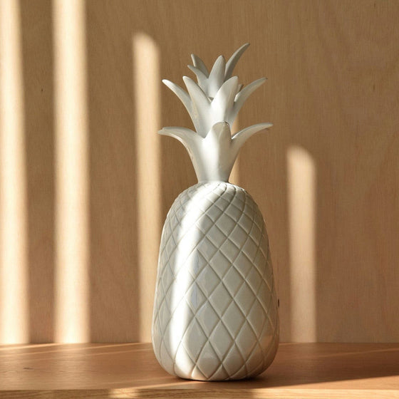 Iluminadas por una luz y sombras suaves, las "Figuras Decorativas en Cerámica Blanca Piña" muestran una artesanía exquisita sobre una mesa de madera. Parte de la colección PIÑA, esta decoración de cerámica texturizada en forma de piña agrega encanto y elegancia a cualquier espacio.