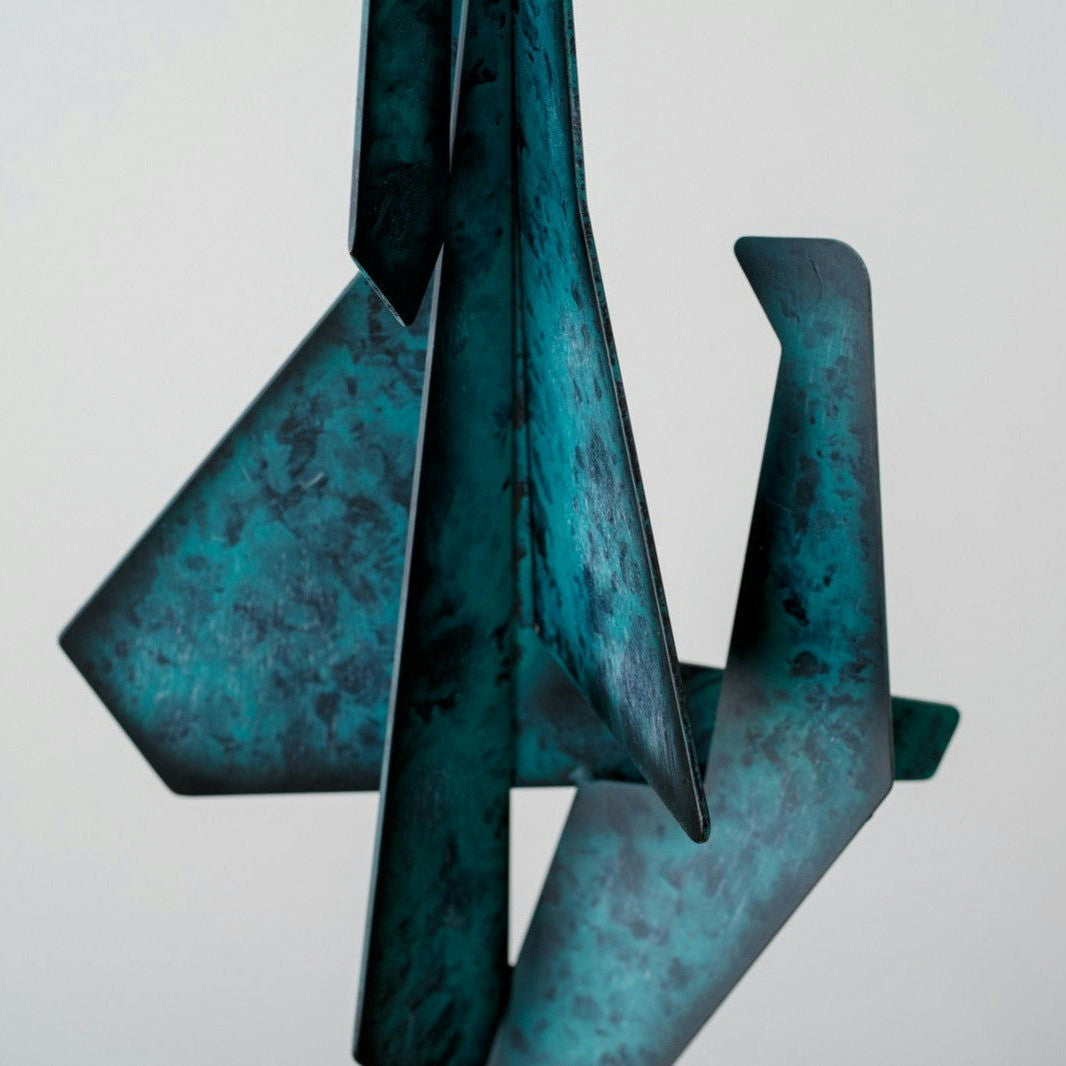 La Figura Decorativa en Metal Verde Abstracción N1 es una escultura abstracta con piezas angulares de metal entrelazadas en acabado turquesa moteado y azul oscuro, suspendida en el aire; captura la esencia del diseño moderno dentro del arte contemporáneo.