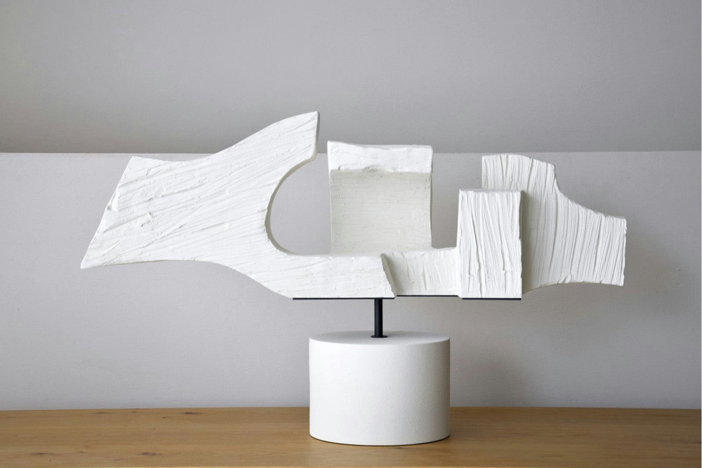 La Figura Decorativa en Cerámica Blanca Hormión es una escultura abstracta versátil con formas angulares y texturizadas montadas sobre una base cilíndrica. Su elegante diseño de cerámica blanca resalta hermosamente sobre una mesa de madera contra un fondo gris neutro.