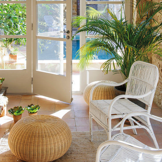 Una habitación iluminada por el sol con sillas de mimbre blanco, el Puff Redondo en Ratán Natural Camel Fritti, plantas en macetas y una puerta de vidrio que exhibe vegetación exterior, exudando elegancia natural.