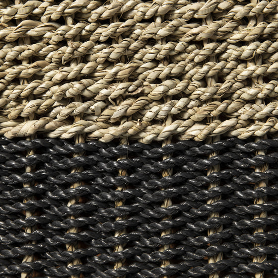 Primer plano de fibras entrelazadas en el Puff de seagrass negro y natural Black/Nat 33x46: fibras de color marrón claro en la parte superior y gris oscuro en la parte inferior, mostrando un tejido intrincado que combina calidad artesanal con diseño moderno.